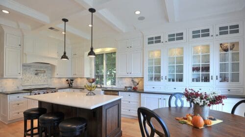 Spotlight on Sherwood: A modern kitchen featuring white cabinetry, an island with black stools, pendant lighting, and a round wooden table adorned with a fall-themed centerpiece for that perfect touch of tradition.