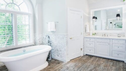 A bright bathroom with a freestanding bathtub, a vanity with double sinks, a large mirror, a white door, and a window with white shutters showcases the perfect HOUZZ Feature: Transitional Bathroom. The floor is wood, and the walls are light-colored.