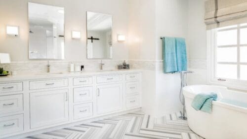 A bright bathroom with white cabinets, two mirrors, a double sink, and wall lights. The Bathroom Renovation: Westlake Transitional design includes a bathtub with blue towels by a large window and a floor featuring a herringbone pattern.