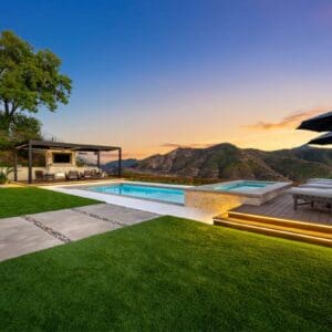 Backyard view with a pool, hot tub, and seating area under a pergola, set against the majestic Sunset Trails mountainous landscape at dusk.