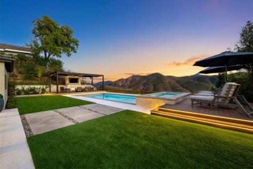 Backyard view with a pool, hot tub, and seating area under a pergola, set against the majestic Sunset Trails mountainous landscape at dusk.