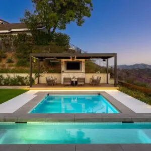 The outdoor pool area, complete with lounge chairs, offers a covered seating space overlooking the enchanting Sunset Trails against a hilly landscape at dusk.