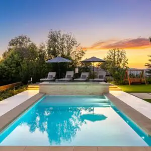 The backyard oasis features a rectangular pool, lounge chairs, and umbrellas, nestled in a lush garden. As the day fades into evening, the vibrant colors of Sunset Trails paint the sky with breathtaking beauty.