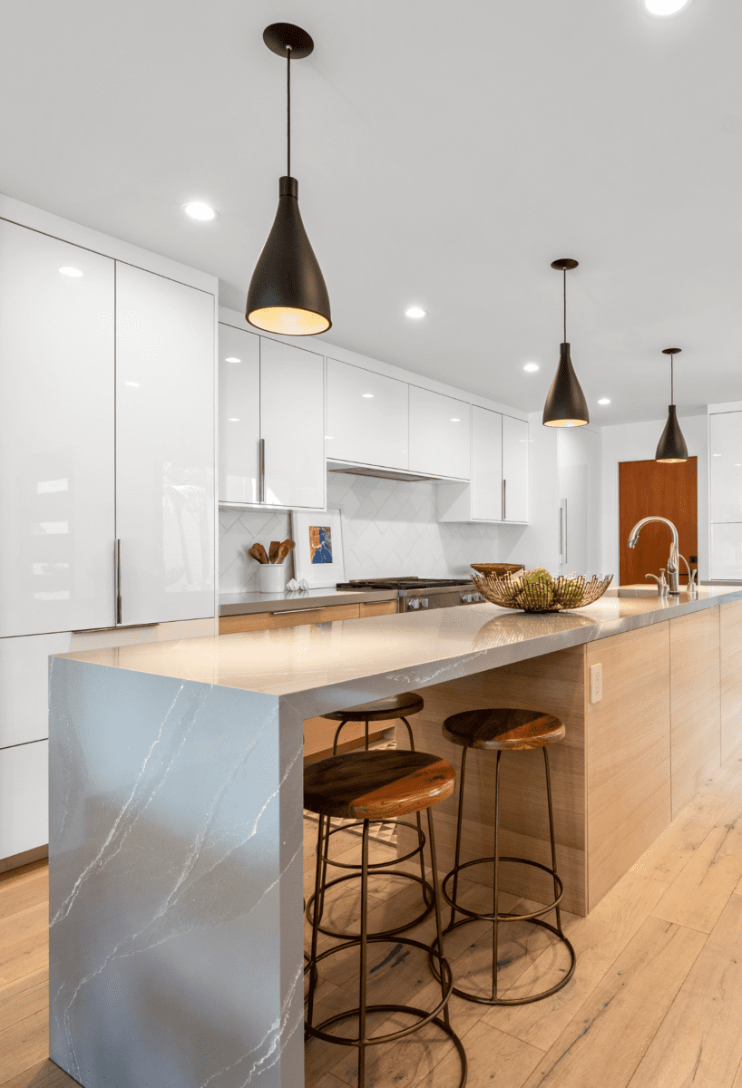 Modern kitchen with white cabinets, marble countertops, and three pendant lights. As part of a contemporary lakeside renovation, a kitchen island with stools, fruit basket, and stainless steel sink is centered in the room.