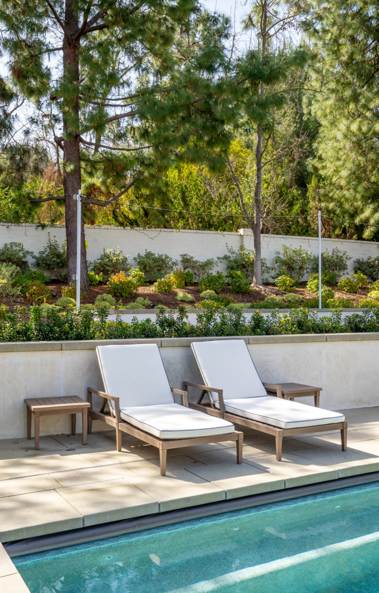 Two wooden lounge chairs with white cushions are placed beside a swimming pool at Westlake Trails. A small wooden table is between the chairs. The area is surrounded by greenery and trees, with a white wall in the background.