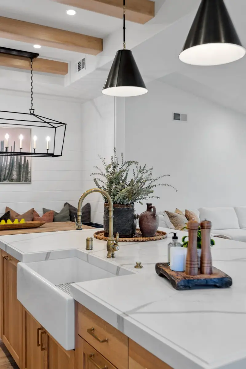 A modern kitchen with a farmhouse sink, wooden cabinetry, pendant lights, and a white countertop. The kitchen opens to a living area with a white couch and decorative pillows.