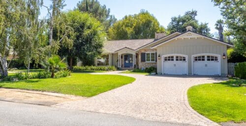 A single-story house with a stone facade, a double garage, and a well-maintained lawn surrounded by trees and shrubs, featuring recent landscaping projects and a paved driveway leading to the front entrance.