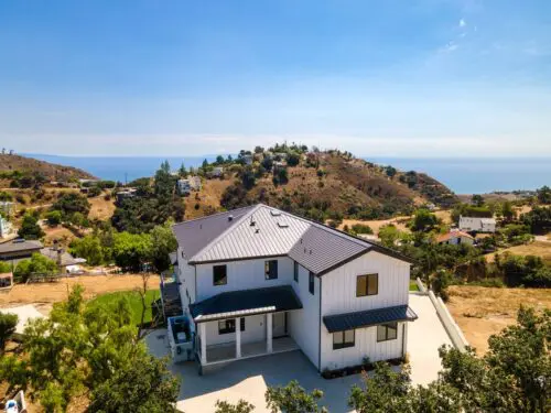 A modern white house with a gray roof is situated in a hilly area with other houses in the background, under a clear blue sky with distant views of the sea.