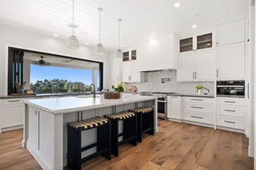 Modern kitchen with white cabinets, large marble island, three black stools, hardwood floors, stainless steel appliances, and an open window view of the scenic Carrita Seaview landscape.