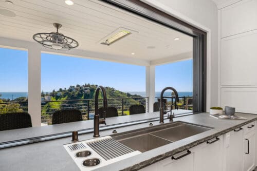 A modern kitchen sink with a large window opens to a spacious patio with outdoor seating, offering a scenic view of the hilly Carrita Seaview landscape and the ocean in the background.