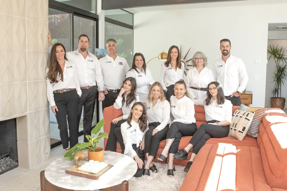 A group of people wearing white shirts and black pants are posing together in a living room. Some are seated on an orange couch while others stand behind it, smiling as if to say "Thank You" for the memories shared.