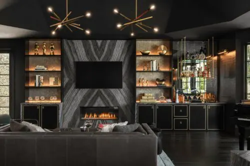 A modern living room exudes sophisticated glamour with a fireplace, wall-mounted TV, built-in shelves adorned with decorative items, a bar area, and stylish lighting fixtures. The dark interior color scheme complements the luxurious furnishings beautifully.