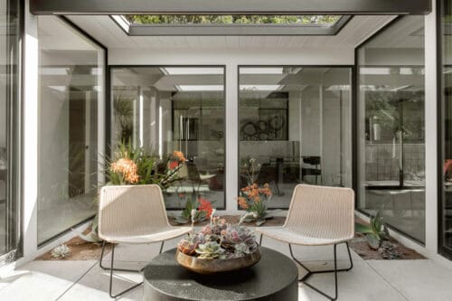 A modern indoor conservatory with large glass panels, skylights, and two beige chairs around a round black table topped with a succulent plant arrangement. The space is subtly accented with touches of Eichler Orange for a warm, inviting ambiance.