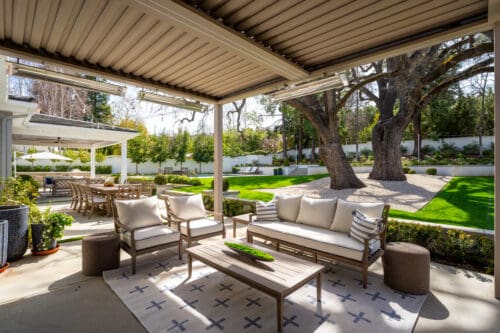 Outdoor patio with modern seating, overhead pergola, and a view of a manicured lawn and garden with large trees. Dining table and chairs are visible in the background, reminiscent of the serene beauty found in Westlake Trails.