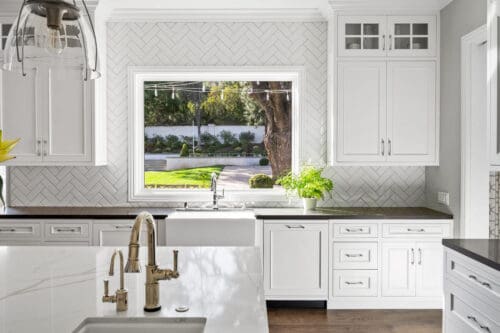 Bright kitchen with white cabinets, a large window above the sink offering a view of the Westlake Trails' lush greenery, and a white herringbone tile backsplash. Marble island with a built-in faucet in the foreground.