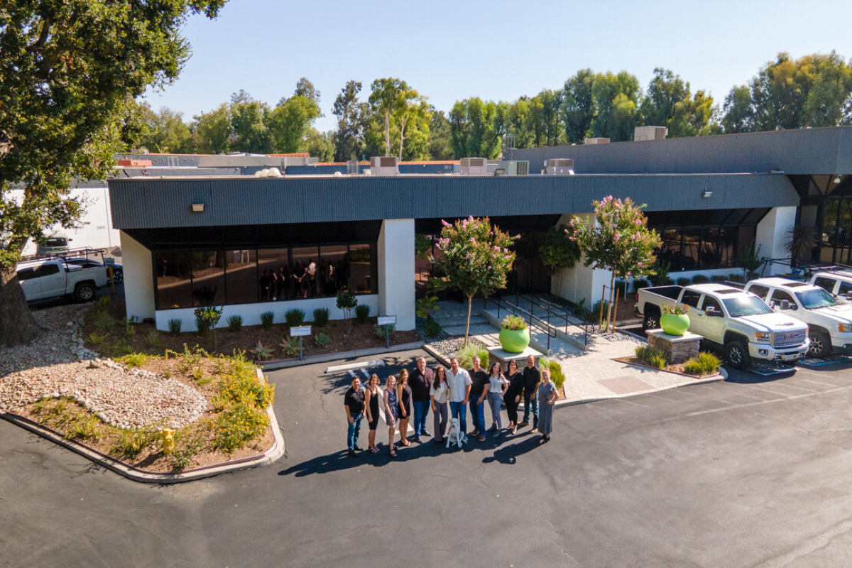 A group of people stands in a parking lot, in front of a modern, one-story building with design-build architecture, surrounded by greenery and several cars.