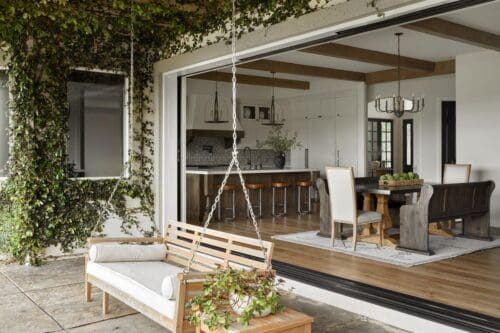 An outdoor patio with a wooden swing bench, adjacent to an open sliding door revealing a modern kitchen and dining area inside a Mediterranean marvel. Ivy climbs the exterior wall.