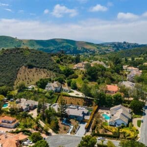 Aerial view of Rustic North Ranch, a suburban neighborhood with scattered homes, greenery, and hills in the background under a partly cloudy sky.