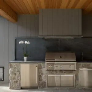 A modern outdoor kitchen in Rustic North Ranch features stainless steel appliances, a stone countertop, and a wooden ceiling. A white orchid in a pot is placed on the counter.