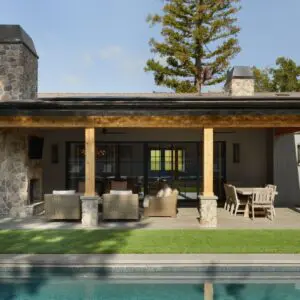 A modern house exterior at Rustic North Ranch features stone columns, wood beams, patio furniture, a dining area, and a pool in the foreground. Trees and a hillside enhance the serene backdrop.