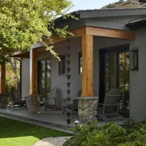 A small, gray house with a stone-accented porch, wooden columns, and two rocking chairs evokes the charm of Rustic North Ranch. The front yard features a manicured lawn, a path, and trees.