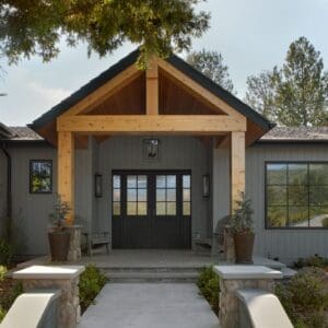 A modern house entrance at Rustic North Ranch features a peaked wooden roof supported by beams, double glass doors, two potted plants, and a stone pathway leading to the front steps. Trees and shrubs surround the entryway, blending contemporary design with rustic charm.