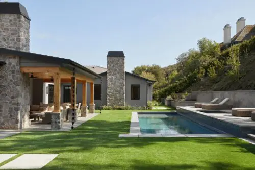 A modern house at Rustic North Ranch boasts a stone facade, a covered patio, a green lawn, and a rectangular swimming pool. Lounge chairs are elegantly positioned on the poolside deck.