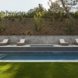 A swimming pool with four lounge chairs and small tables arranged on the pool deck. In the backdrop, the lush greenery of Rustic North Ranch adds a charming, natural touch with its hedge and trees.