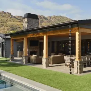 A modern house with a wooden patio area, outdoor seating, and a stone fireplace is set against the rustic North Ranch mountains, next to a swimming pool surrounded by a well-manicured lawn.