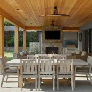 Experience the charm of Rustic North Ranch on this covered outdoor patio featuring a wooden ceiling, stone fireplace, and large dining table with chairs. The seating area includes cushioned chairs and a sofa under ceiling fans, seamlessly opening up to a beautifully landscaped yard.