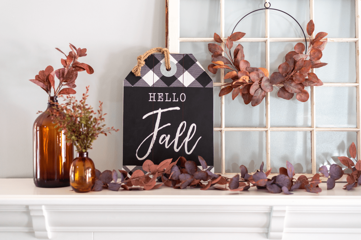 A fall-themed display featuring amber bottles with autumn foliage, a "Hello Fall" sign, and a wreath of red leaves hanging on a window frame blends seasonal decor seamlessly into your home.