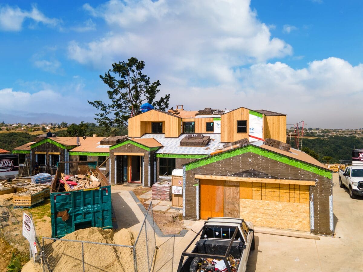 A large house under construction, part of a fire rebuild effort, showcases visible wooden framing and a partially completed roof. Construction materials and vehicles are actively present on site.