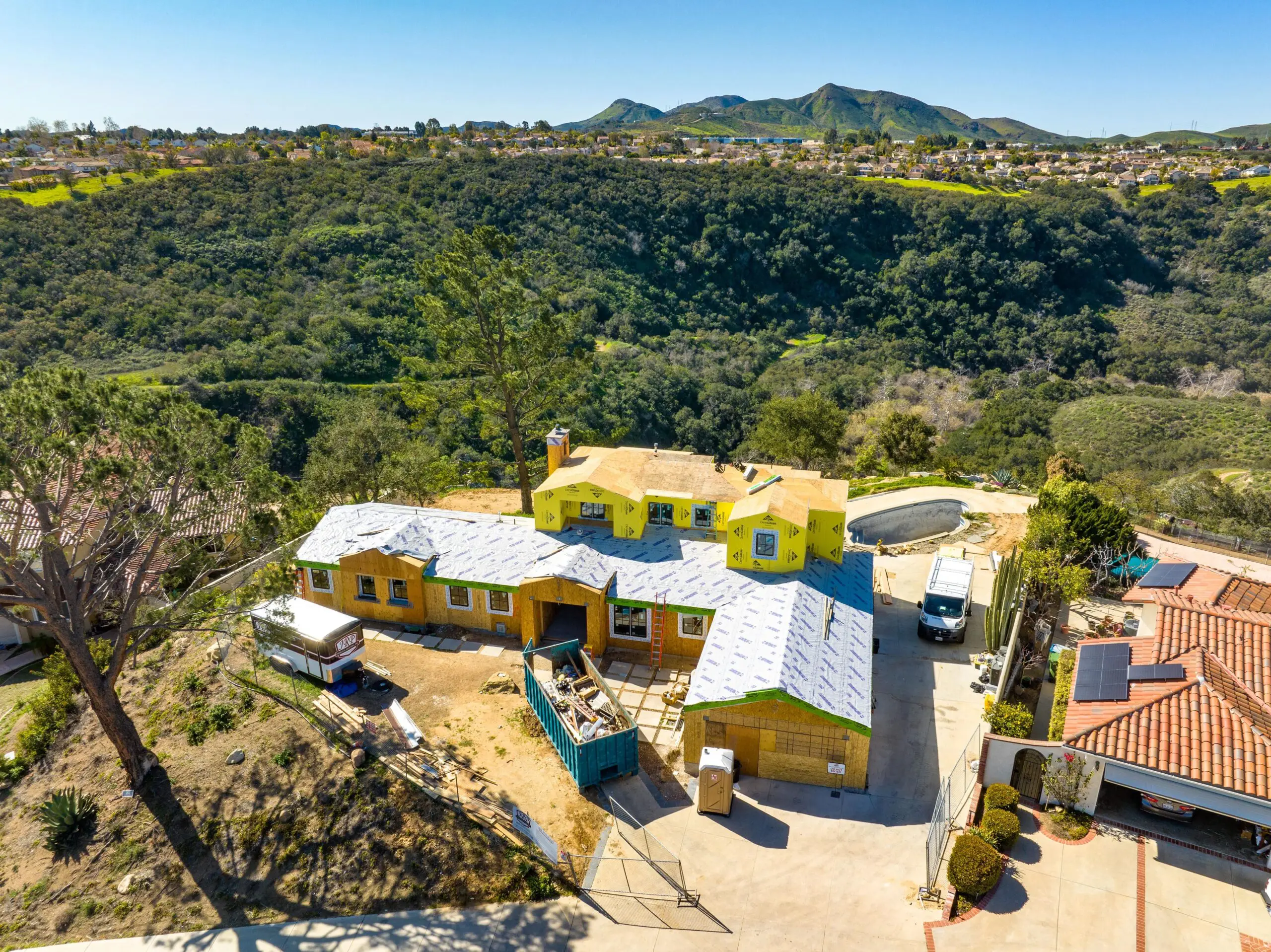 Aerial view of a hilltop house under construction, part of a fire rebuild effort, with a partially completed roof. Its surrounded by trees and overlooks a valley and distant hills.