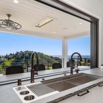 A modern kitchen sink with a large window opens to a spacious patio with outdoor seating, offering a scenic view of the hilly Carrita Seaview landscape and the ocean in the background.