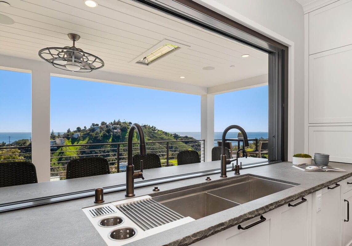 A modern kitchen sink with a large window opens to a spacious patio with outdoor seating, offering a scenic view of the hilly Carrita Seaview landscape and the ocean in the background.