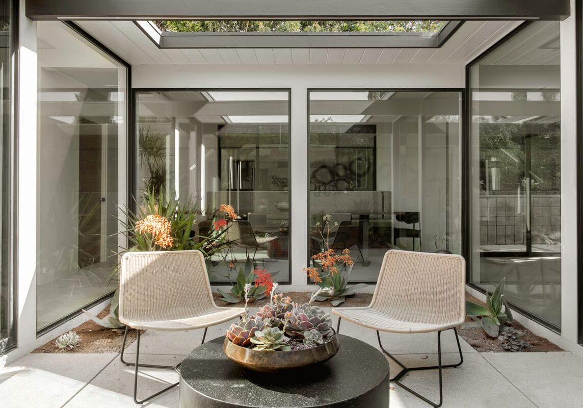 A modern indoor conservatory with large glass panels, skylights, and two beige chairs around a round black table topped with a succulent plant arrangement. The space is subtly accented with touches of Eichler Orange for a warm, inviting ambiance.