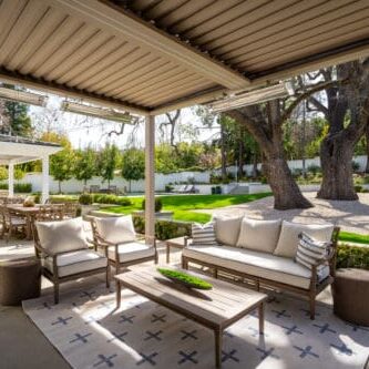 Outdoor patio with modern seating, overhead pergola, and a view of a manicured lawn and garden with large trees. Dining table and chairs are visible in the background, reminiscent of the serene beauty found in Westlake Trails.