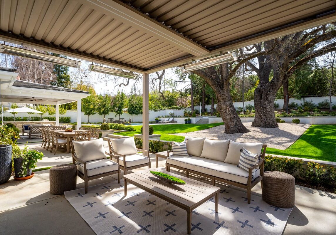 Outdoor patio with modern seating, overhead pergola, and a view of a manicured lawn and garden with large trees. Dining table and chairs are visible in the background, reminiscent of the serene beauty found in Westlake Trails.