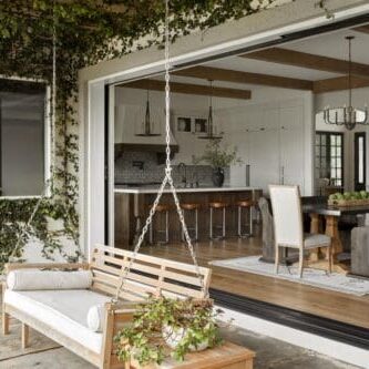 An outdoor patio with a wooden swing bench, adjacent to an open sliding door revealing a modern kitchen and dining area inside a Mediterranean marvel. Ivy climbs the exterior wall.