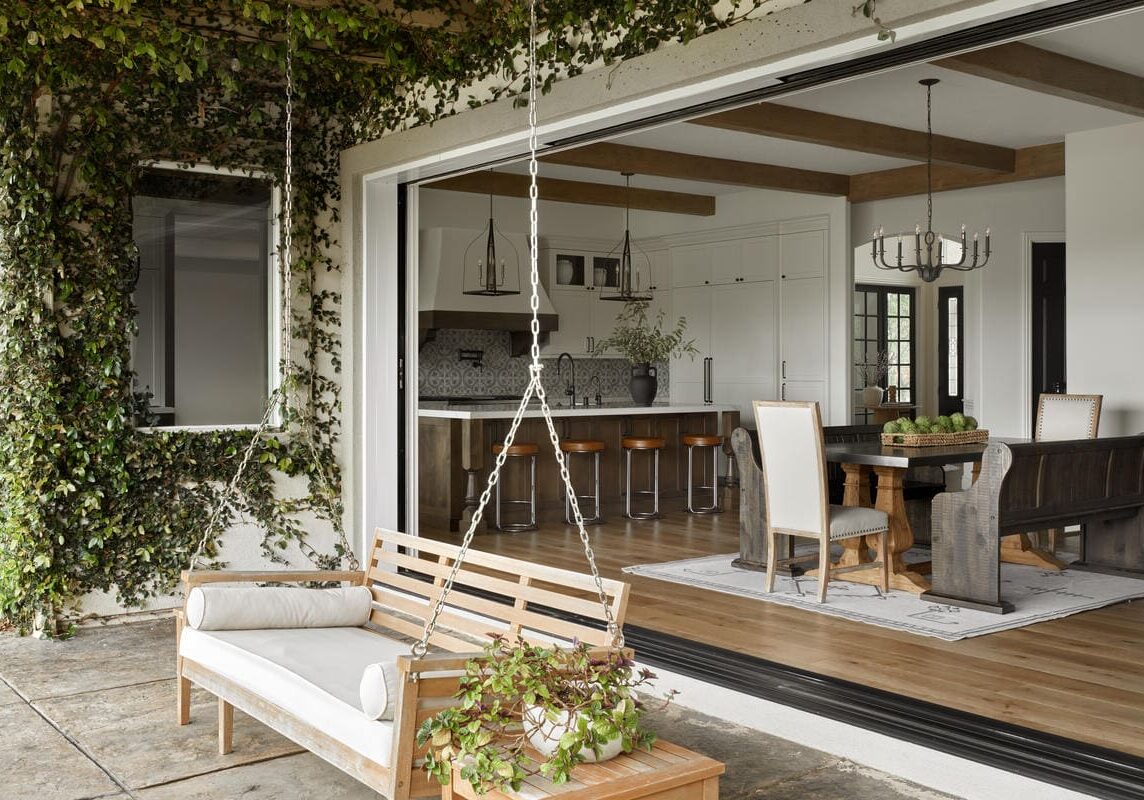 An outdoor patio with a wooden swing bench, adjacent to an open sliding door revealing a modern kitchen and dining area inside a Mediterranean marvel. Ivy climbs the exterior wall.