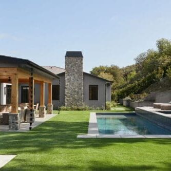 A modern house at Rustic North Ranch boasts a stone facade, a covered patio, a green lawn, and a rectangular swimming pool. Lounge chairs are elegantly positioned on the poolside deck.