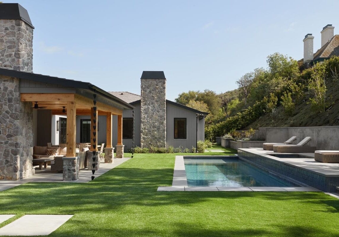 A modern house at Rustic North Ranch boasts a stone facade, a covered patio, a green lawn, and a rectangular swimming pool. Lounge chairs are elegantly positioned on the poolside deck.