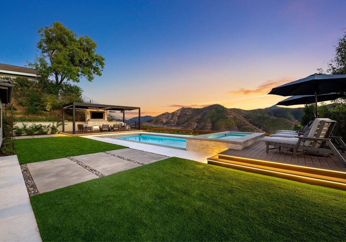 Backyard view with a pool, hot tub, and seating area under a pergola, set against the majestic Sunset Trails mountainous landscape at dusk.